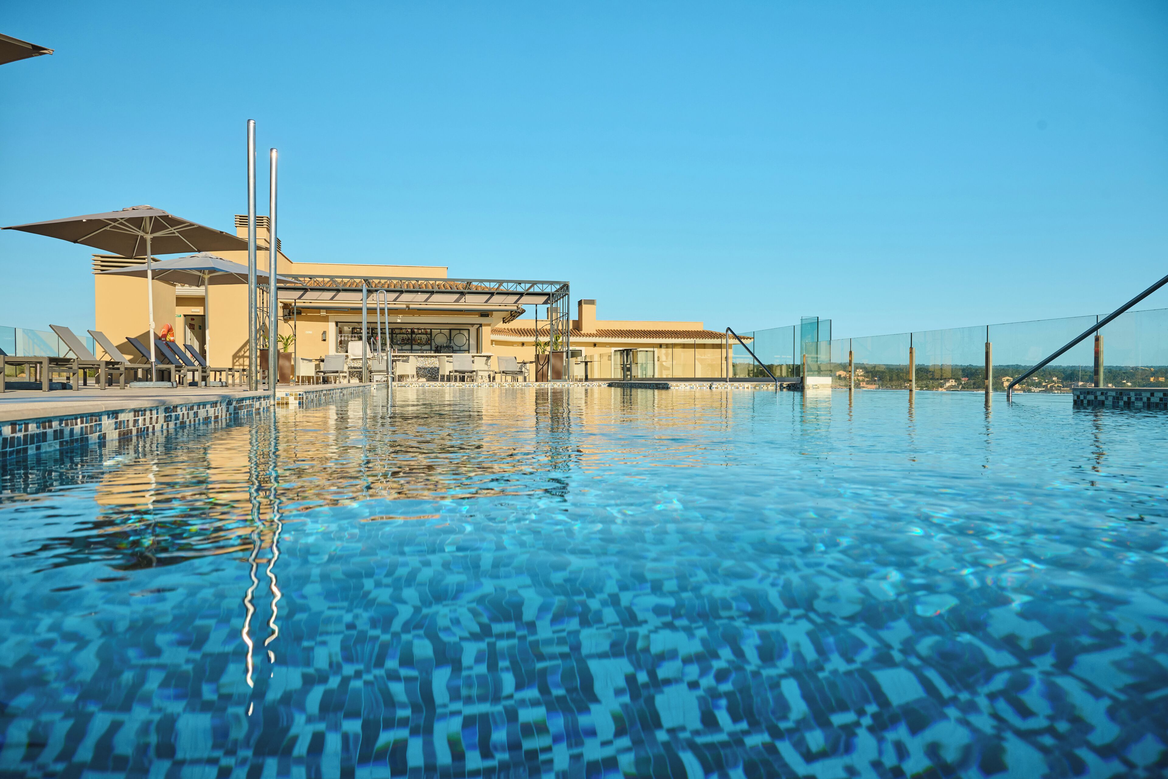 Una piscina con agua azul cristalina, rodeada de sillas de descanso y sombrillas, y un edificio con diseño moderno al fondo.