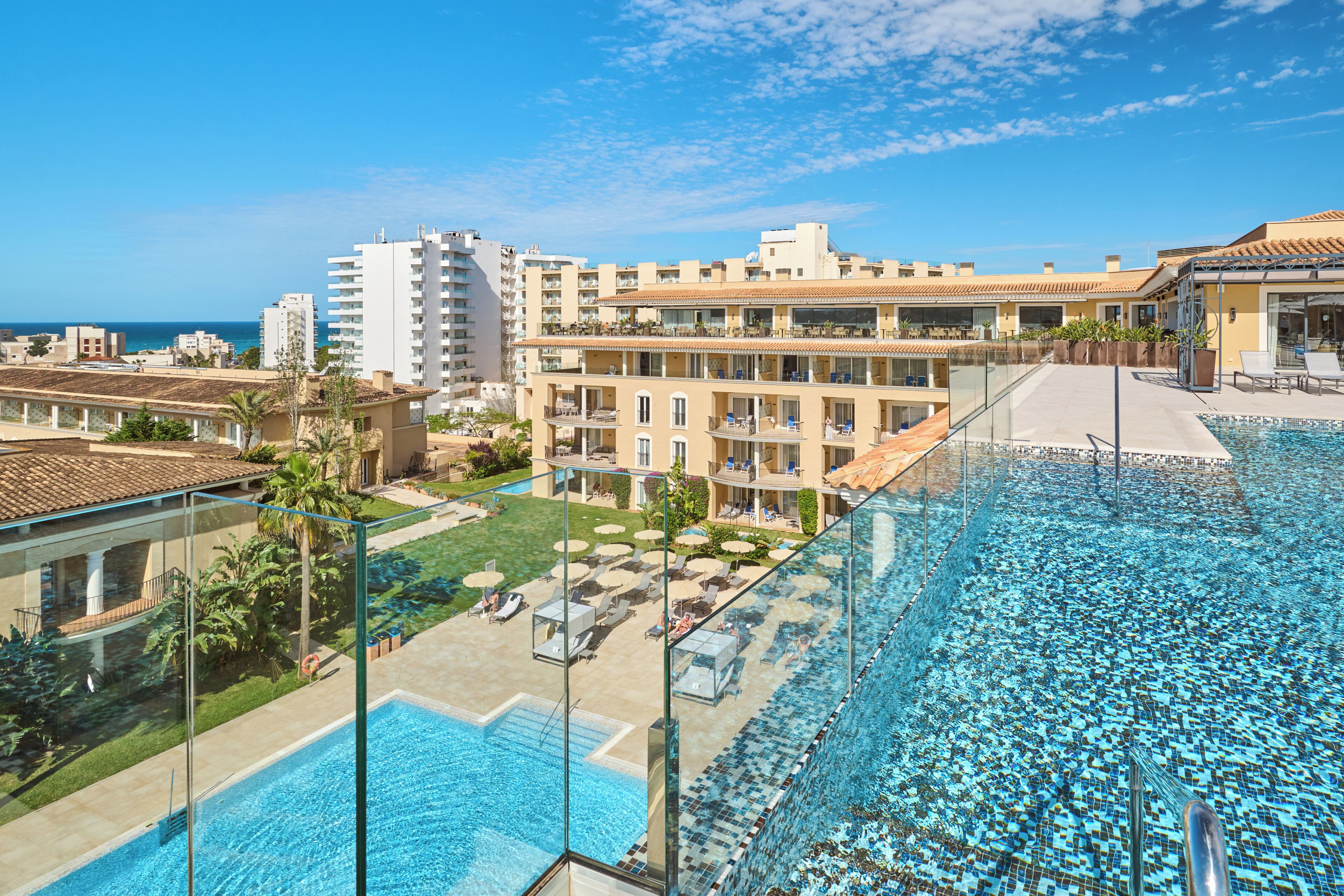 A rooftop pool with clear blue water, surrounded by lounge chairs and umbrellas, overlooking a cityscape with buildings and a view of the sea.