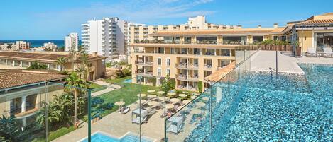 A rooftop pool with clear blue water, surrounded by lounge chairs and umbrellas, overlooking a cityscape with buildings and a view of the sea.