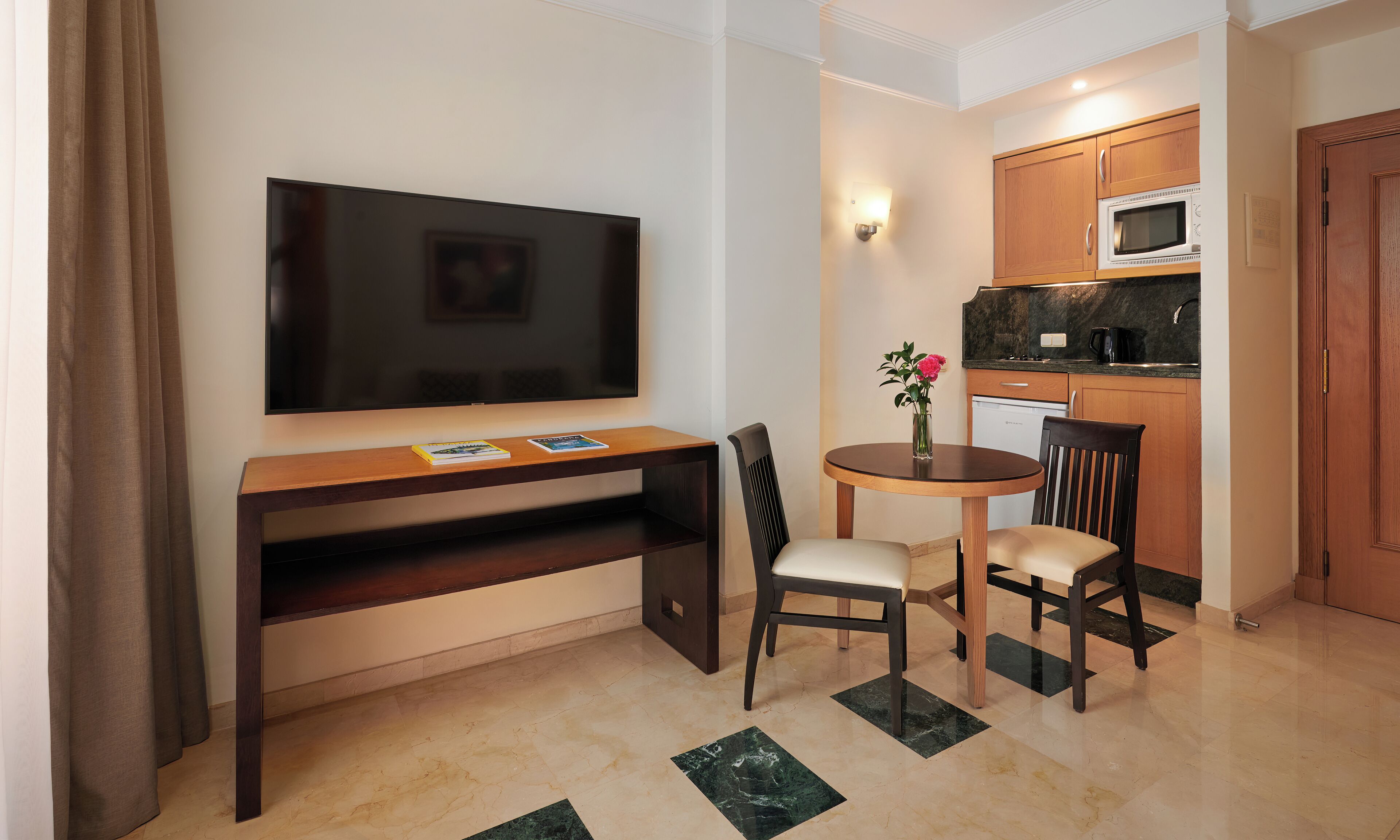 Un espacio habitable compacto con televisión de pantalla plana, una pequeña mesa de comedor con dos sillas, un área de cocina con microondas y un armario de madera.