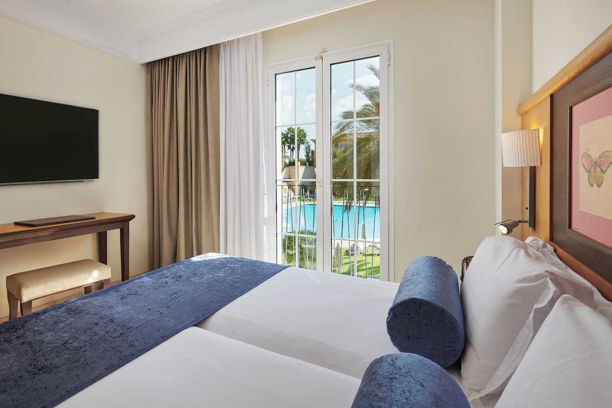 A hotel room with a bed, a desk, a chair, a television, a window with a view of a pool, and a lamp.