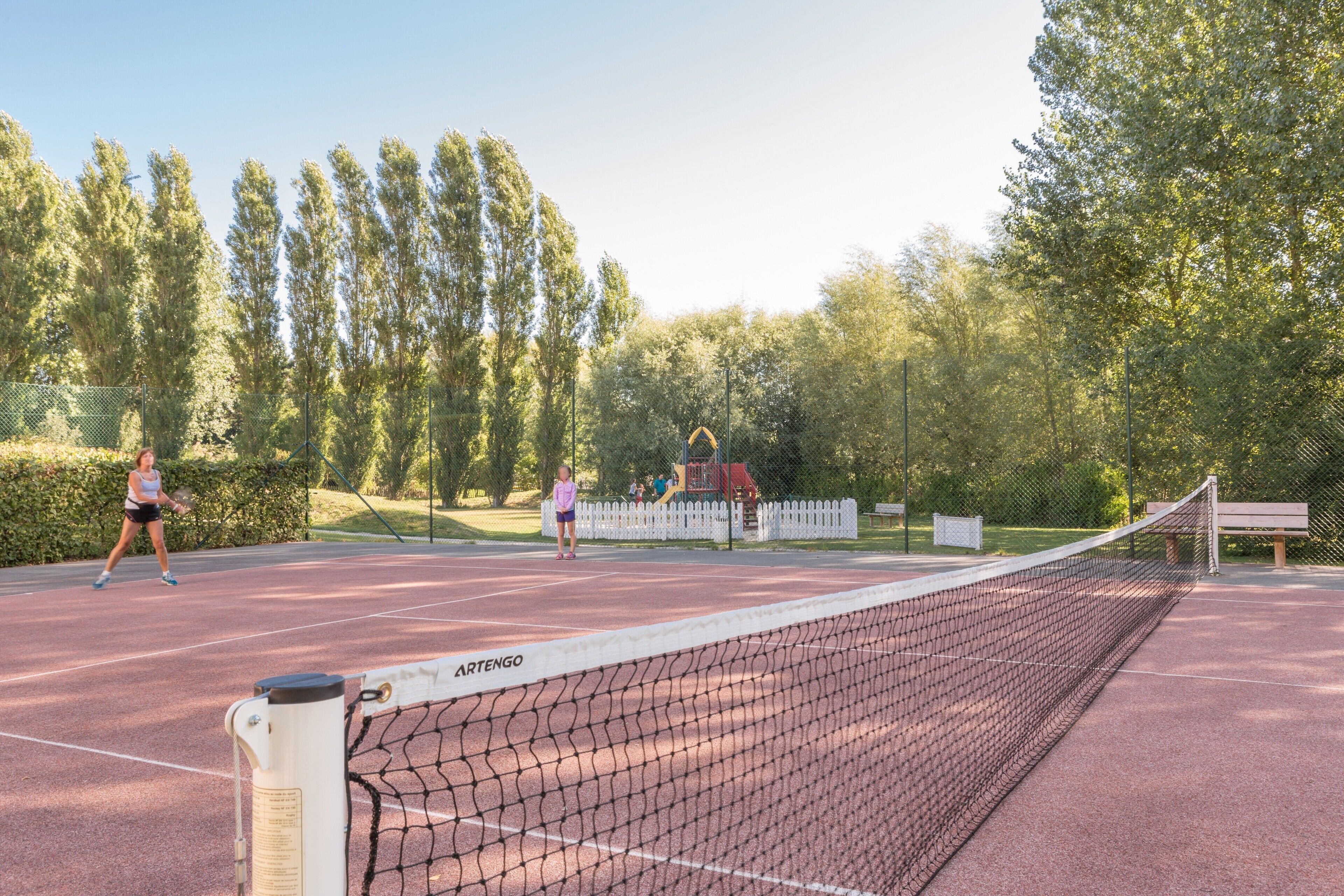 A tennis court with a net labeled "ARTENGO," surrounded by trees and a playground.