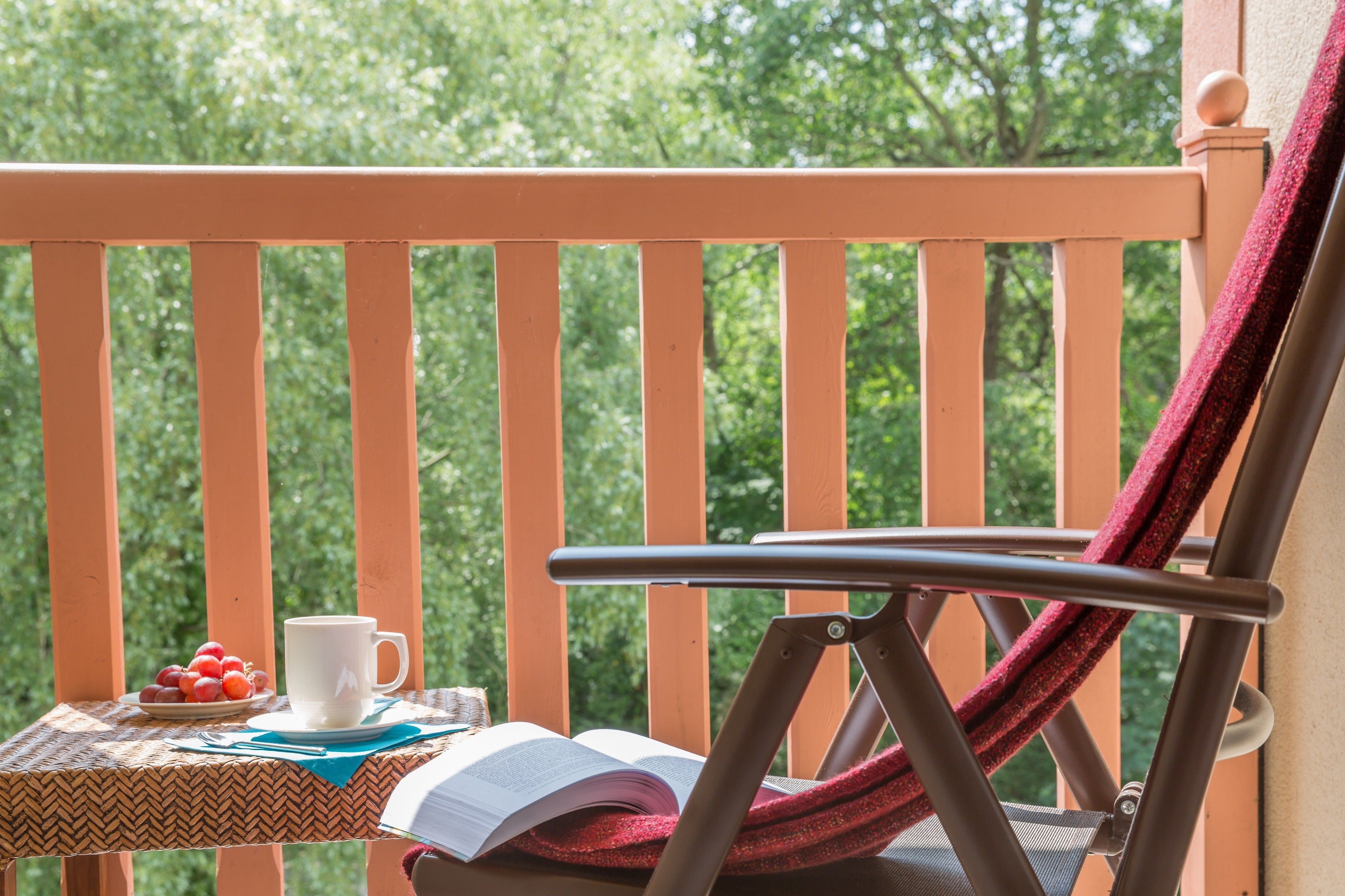 A wooden balcony with a woven table, a chair with a red fabric, a white mug, and a plate of fruit.