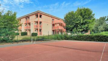 Un court de tennis avec un filet, entouré d’une clôture, et un bâtiment avec des balcons en arrière-plan.