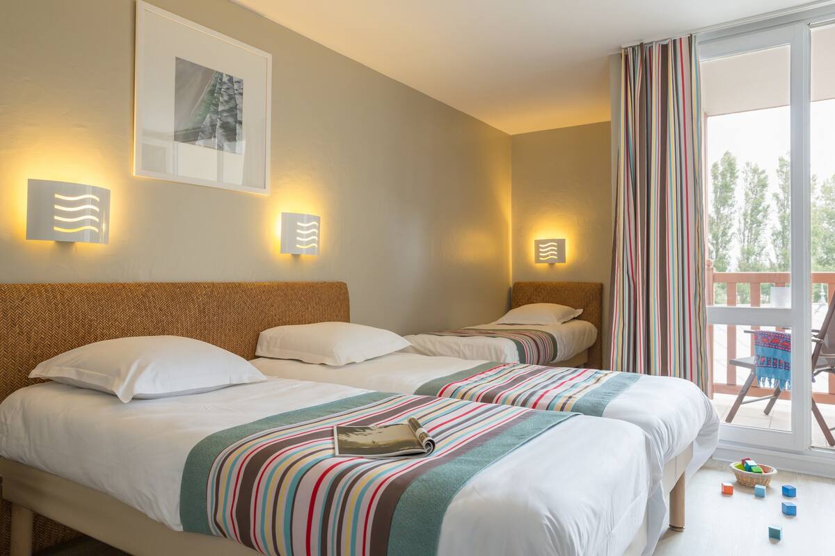 A hotel room with two beds, striped bedding, wall-mounted lights, a framed picture, and a balcony with a view.