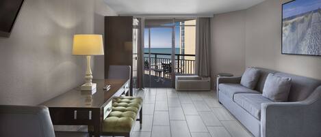 A hotel room with a balcony, a sofa, a dining table with a lamp, and a TV.