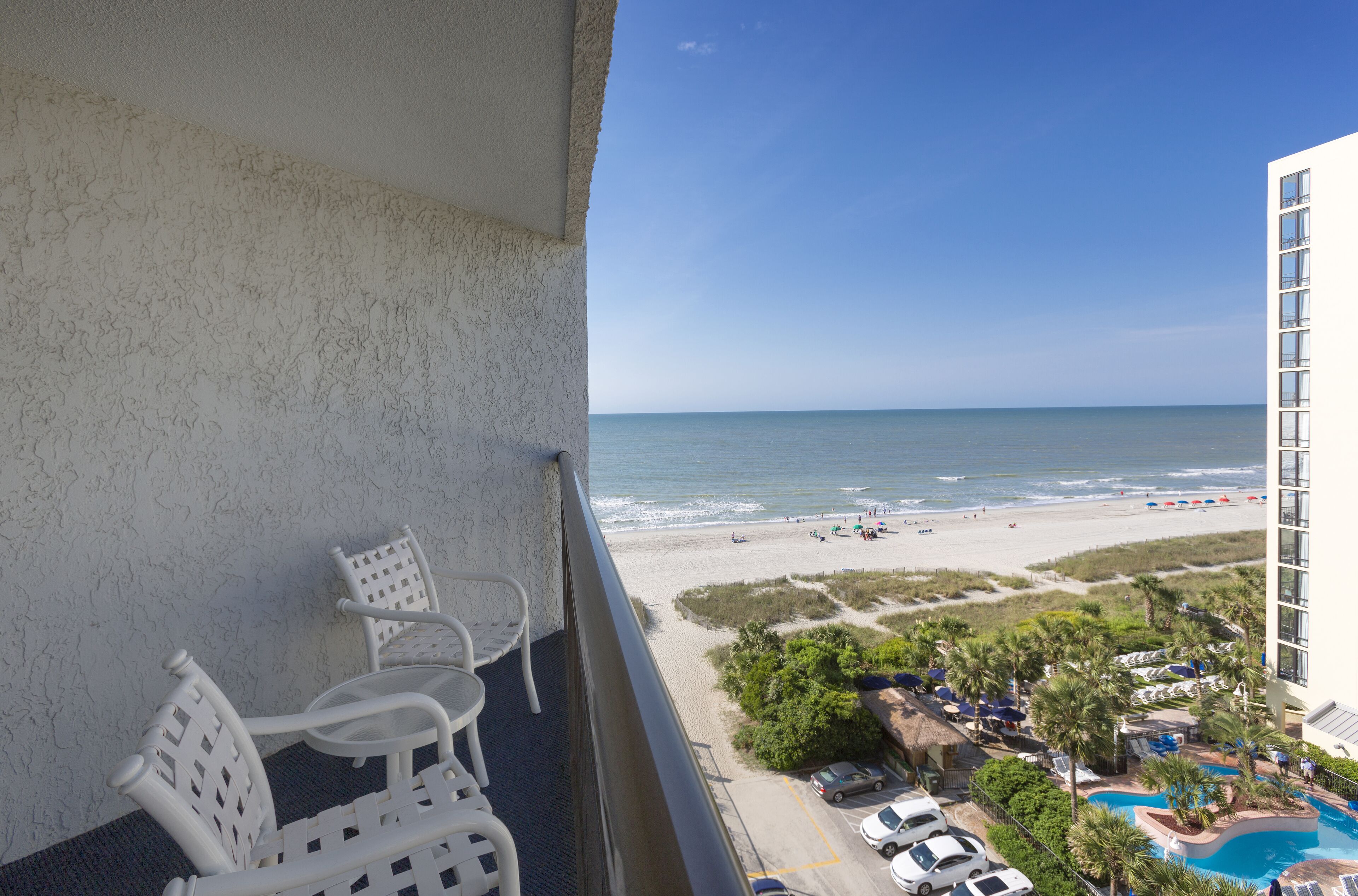 A balcony with white chairs overlooking a beach, a pool, and a building.