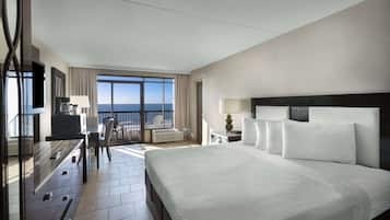 A hotel room with a large bed, a desk with a chair, a TV, and a balcony with a view of the ocean.