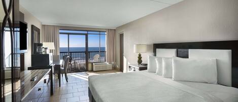 A hotel room with a large bed, a desk with a chair, a TV, and a balcony with a view of the ocean.