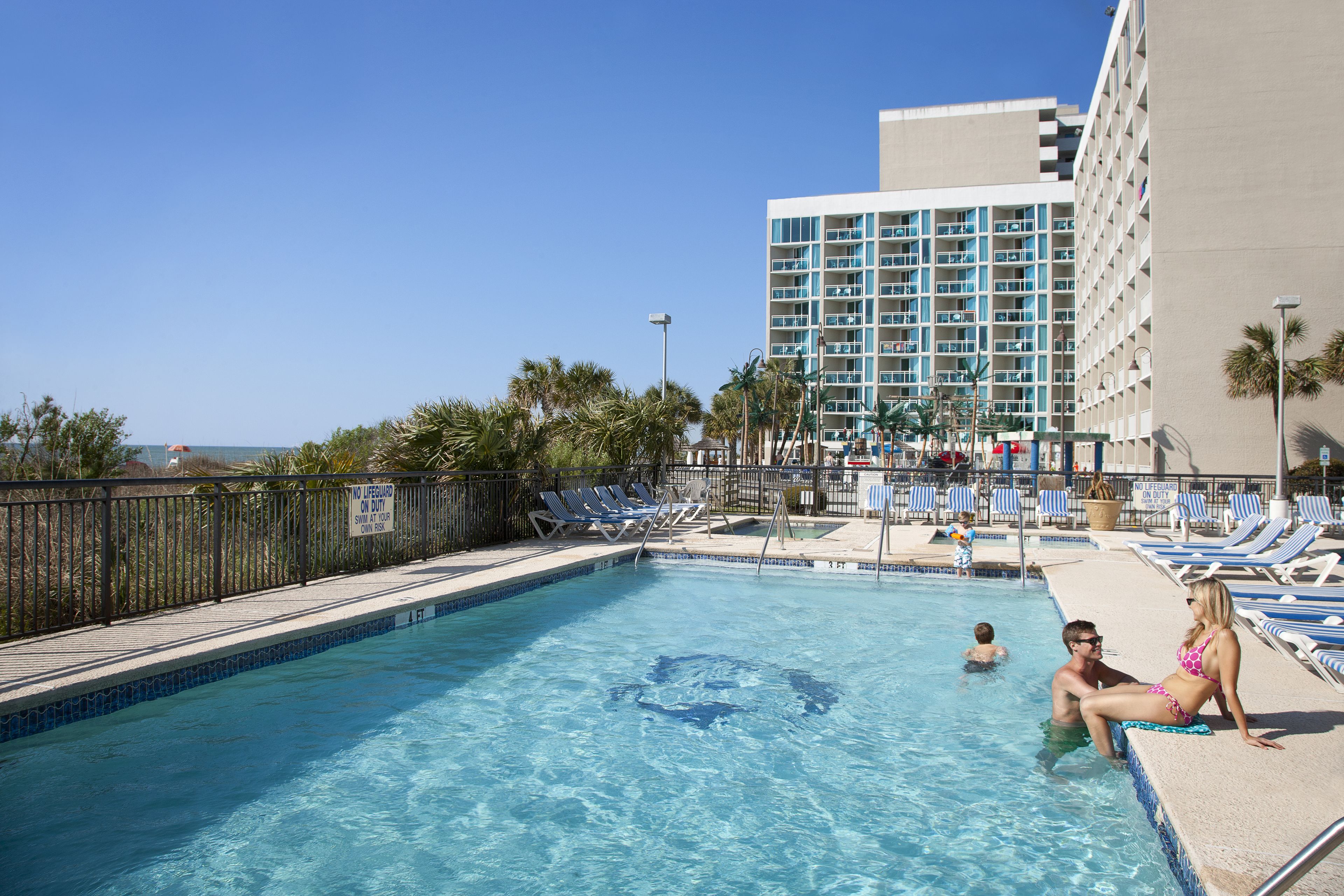 A swimming pool with lounge chairs, a lifeguard tower, and a high-rise building in the background.