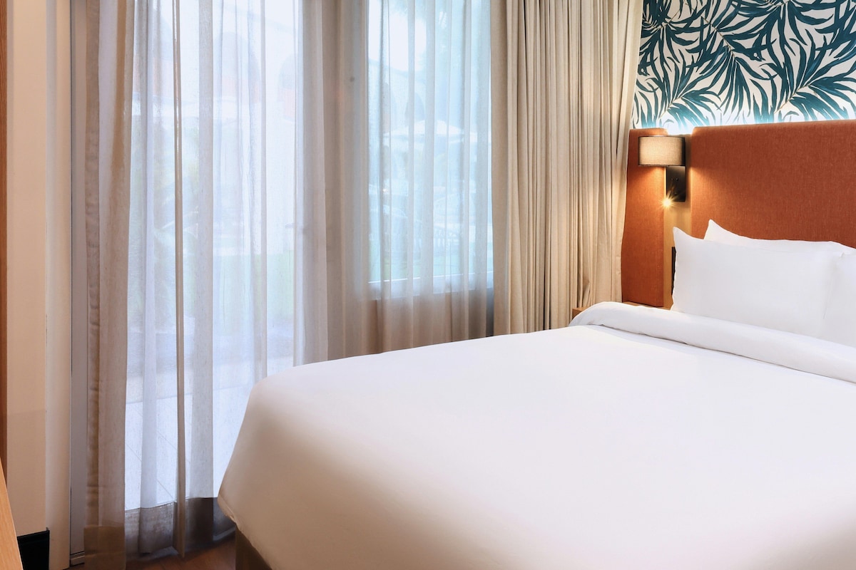 Quarto de hotel com uma cama grande, cabeceira com luminária e cortinas leves.