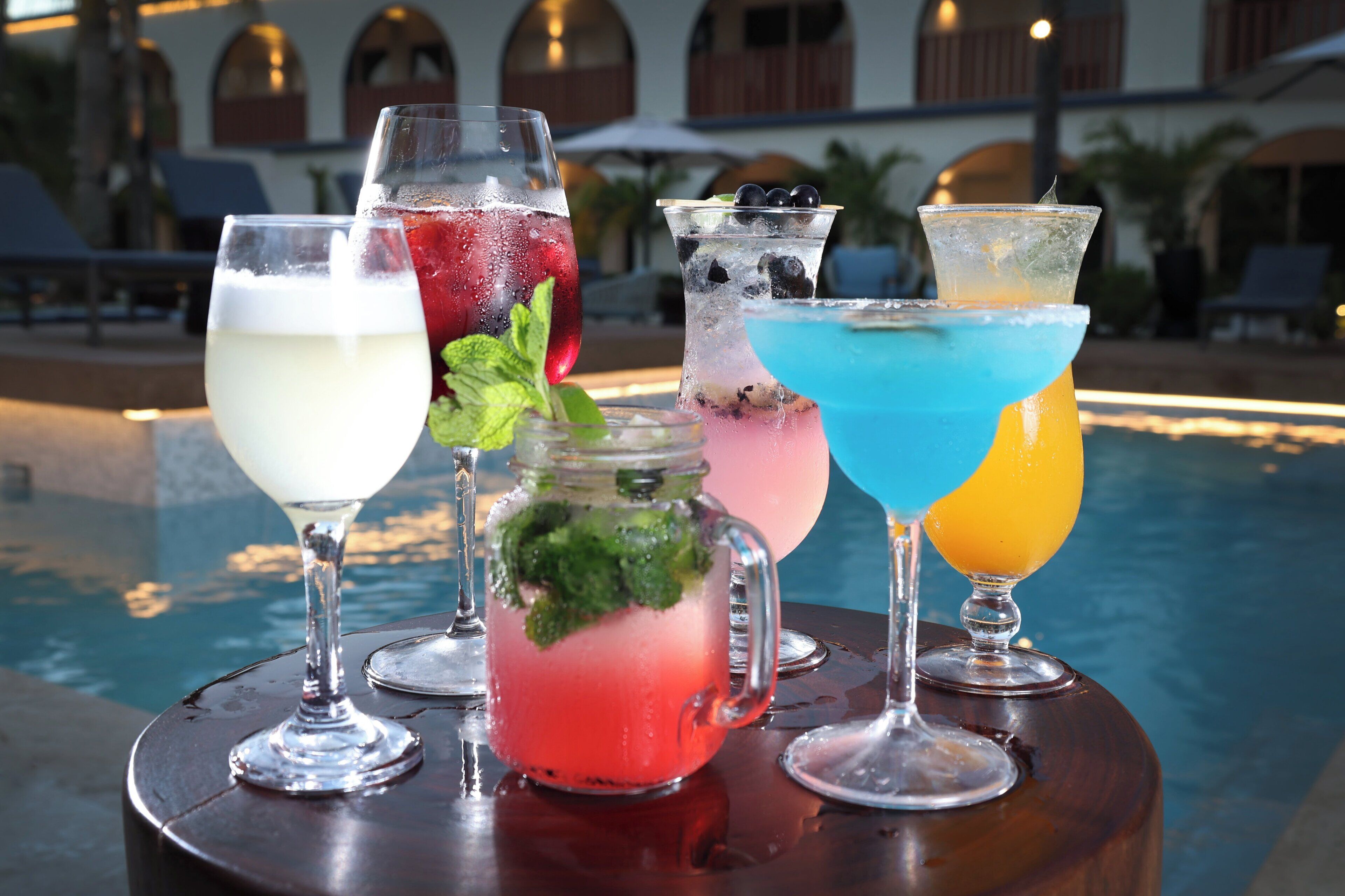 A selection of drinks on a table by a poolside, with a building and lounge chairs in the background.