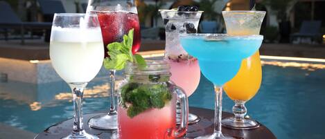 A selection of drinks on a table by a poolside, with a building and lounge chairs in the background.