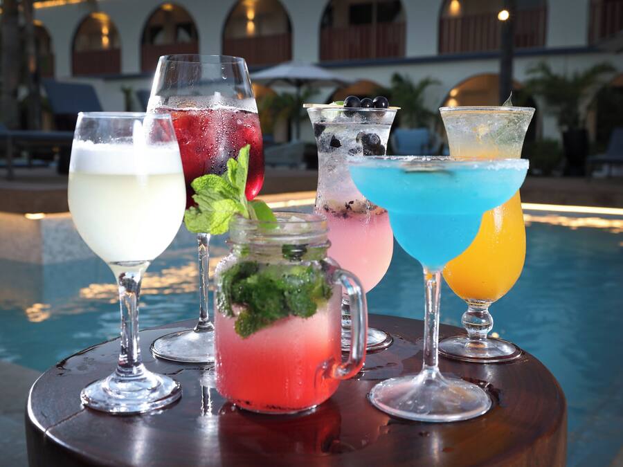 Variedad de bebidas sobre una mesa junto a una piscina, con un edificio y sillones de descanso al fondo.