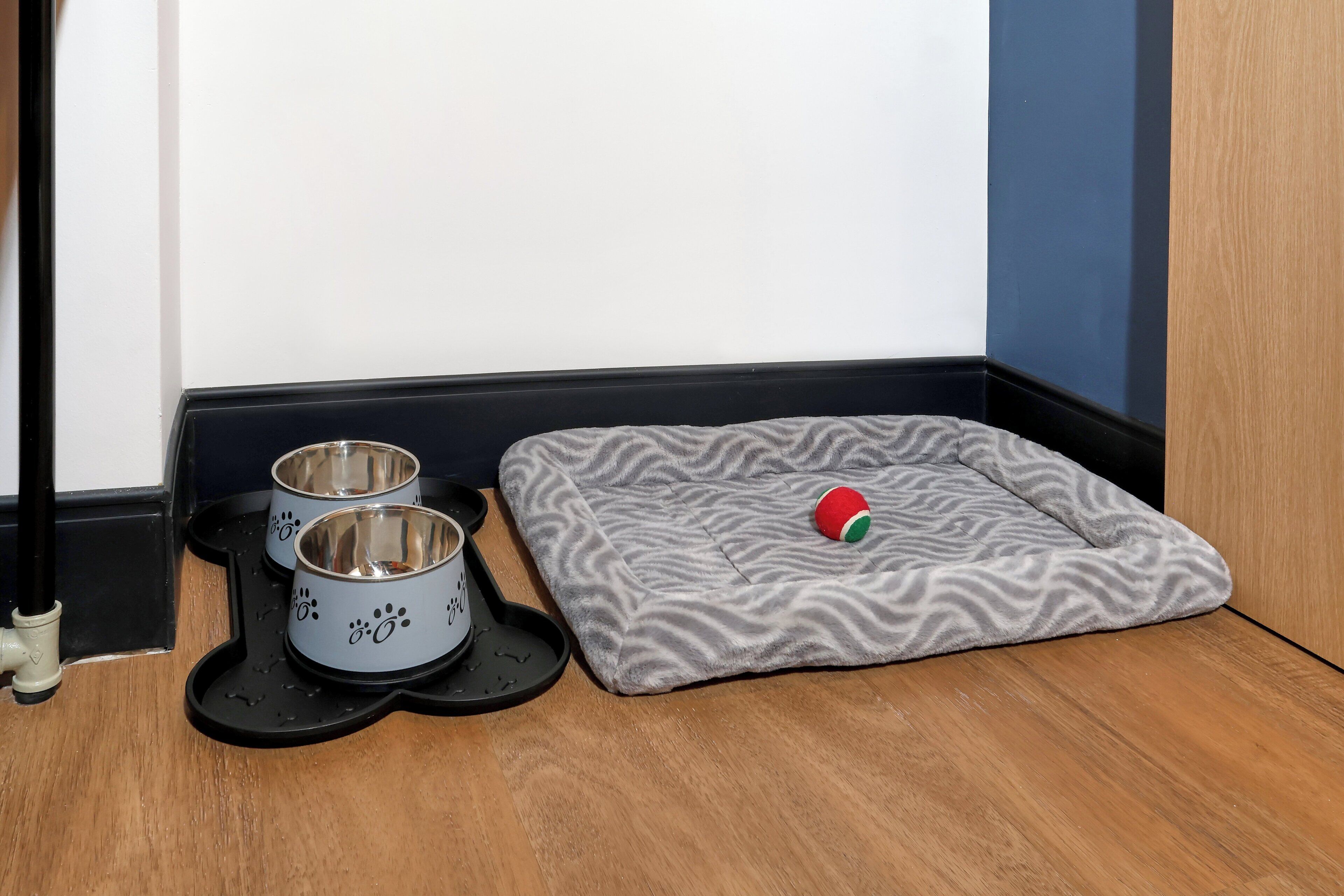Un panier pour animal de compagnie avec un revêtement à motifs et deux gamelles posées sur un plancher en bois.