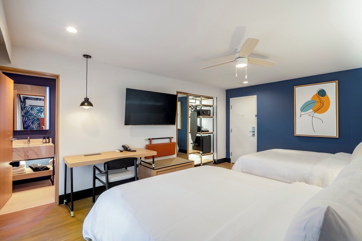 Habitación de hotel con cama, escritorio, silla, televisor, armario y ventilador de techo.