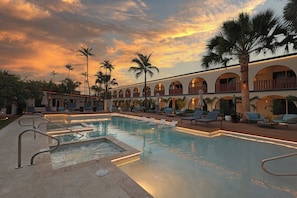 Zona de piscina con sillones de descanso, palmeras y un edificio con ventanales arqueados.