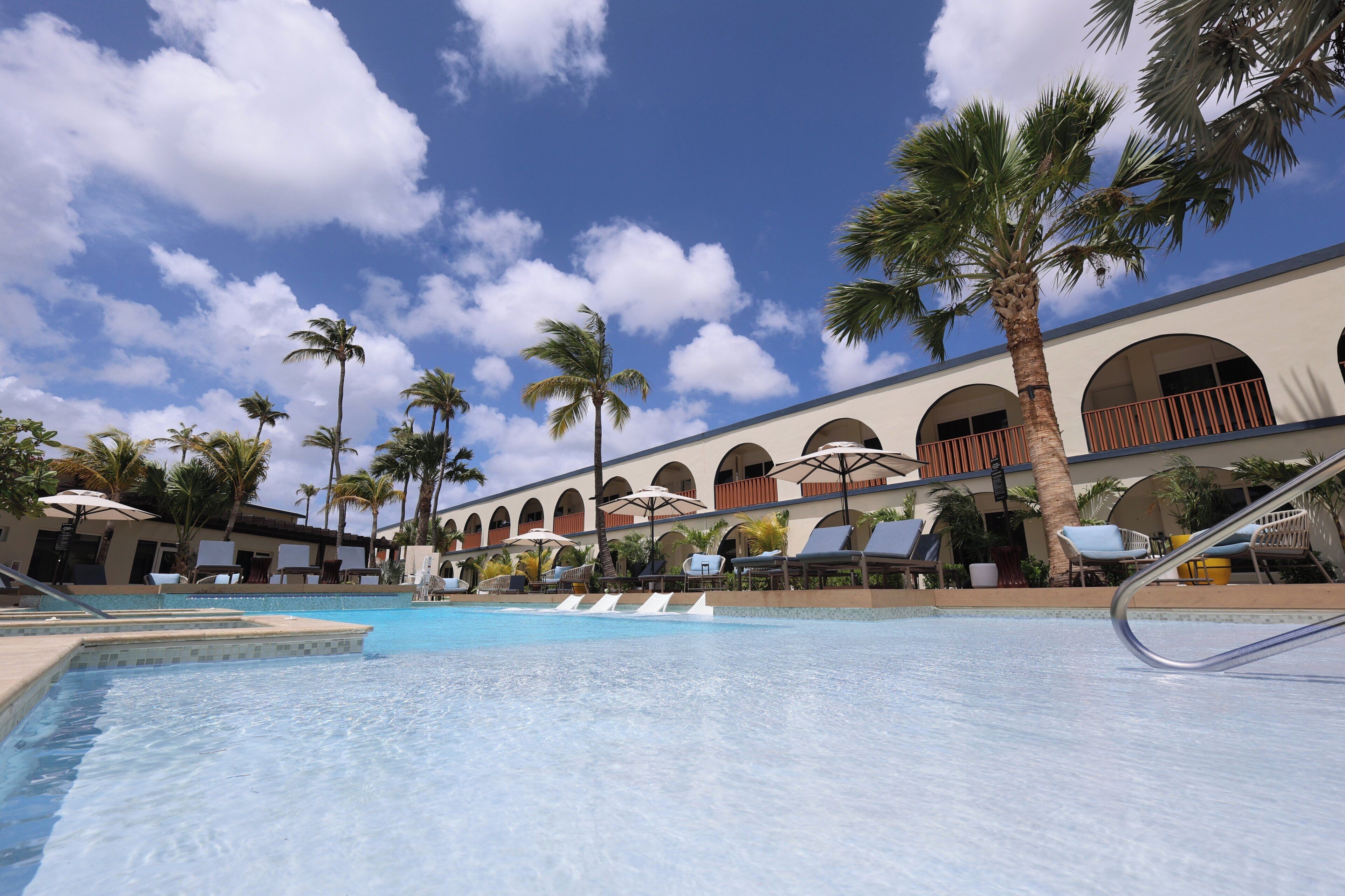 A swimming pool with lounge chairs and umbrellas, surrounded by palm trees and a building with arched windows.