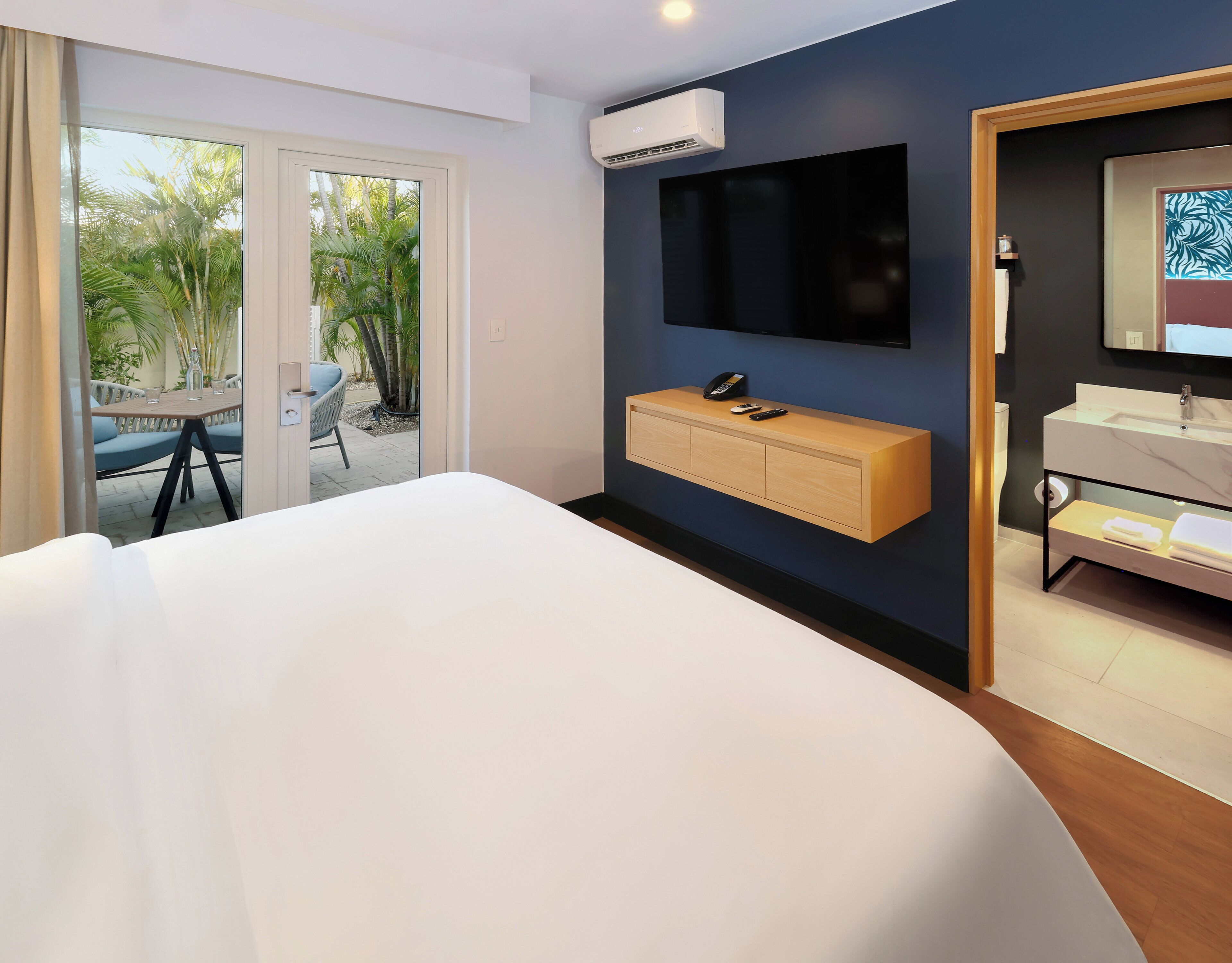 Ein Hotelzimmer mit Bett, Wandfernseher, Holzkonsolentisch, Badezimmer mit Waschbecken und Spiegel sowie eine Glastür, die zu einem Außenbereich mit Pflanzen führt.