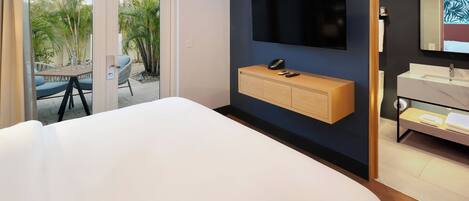Ein Hotelzimmer mit Bett, Wandfernseher, Holzkonsolentisch, Badezimmer mit Waschbecken und Spiegel sowie eine Glastür, die zu einem Außenbereich mit Pflanzen führt.