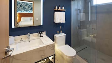 A modern bathroom with a marble sink, a toilet, a glass shower enclosure, and a mirror reflecting a bedroom.
