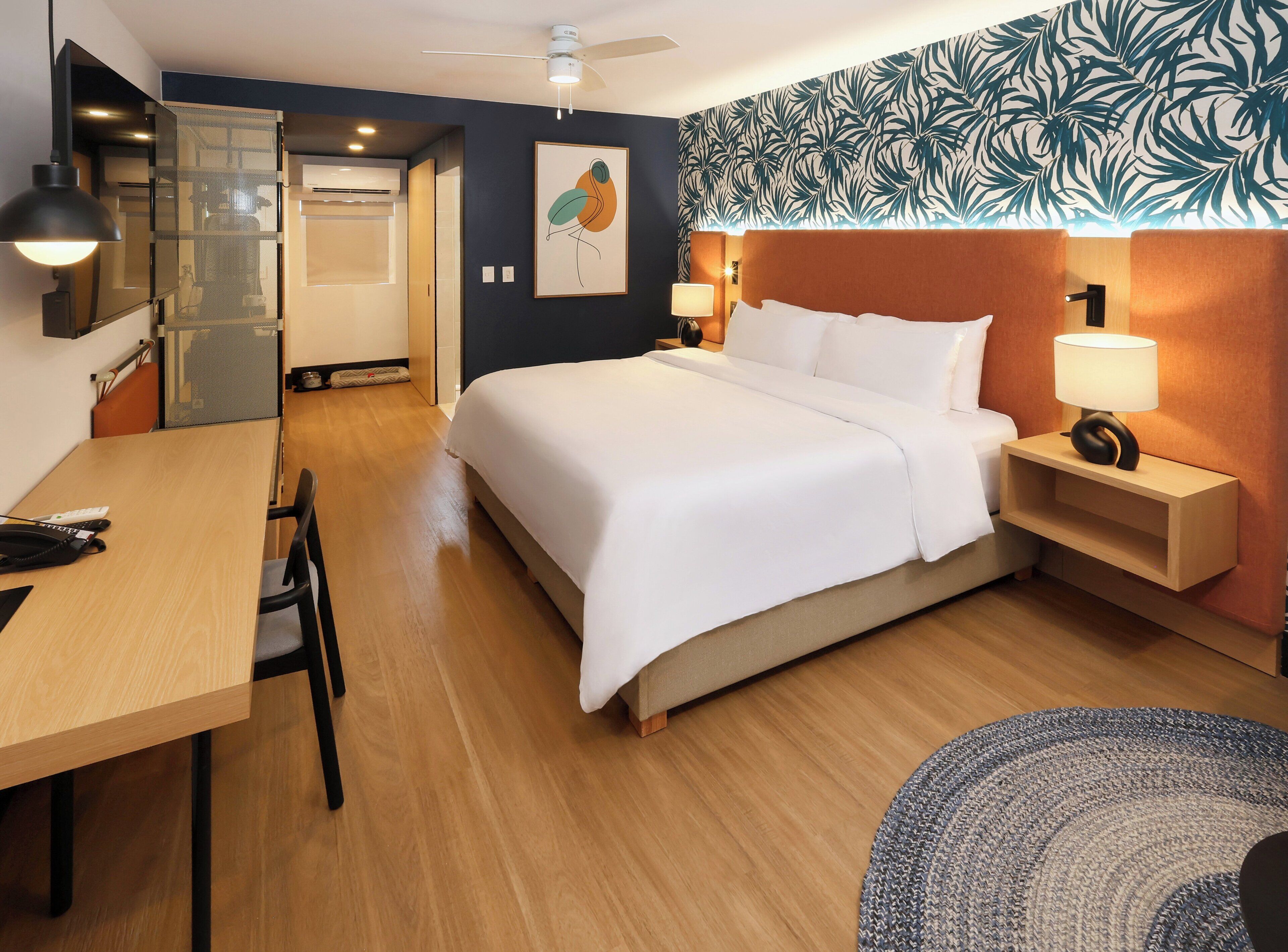 A hotel room with a bed, bedside tables, a desk, a chair, a ceiling fan, and a wall with a leaf pattern.