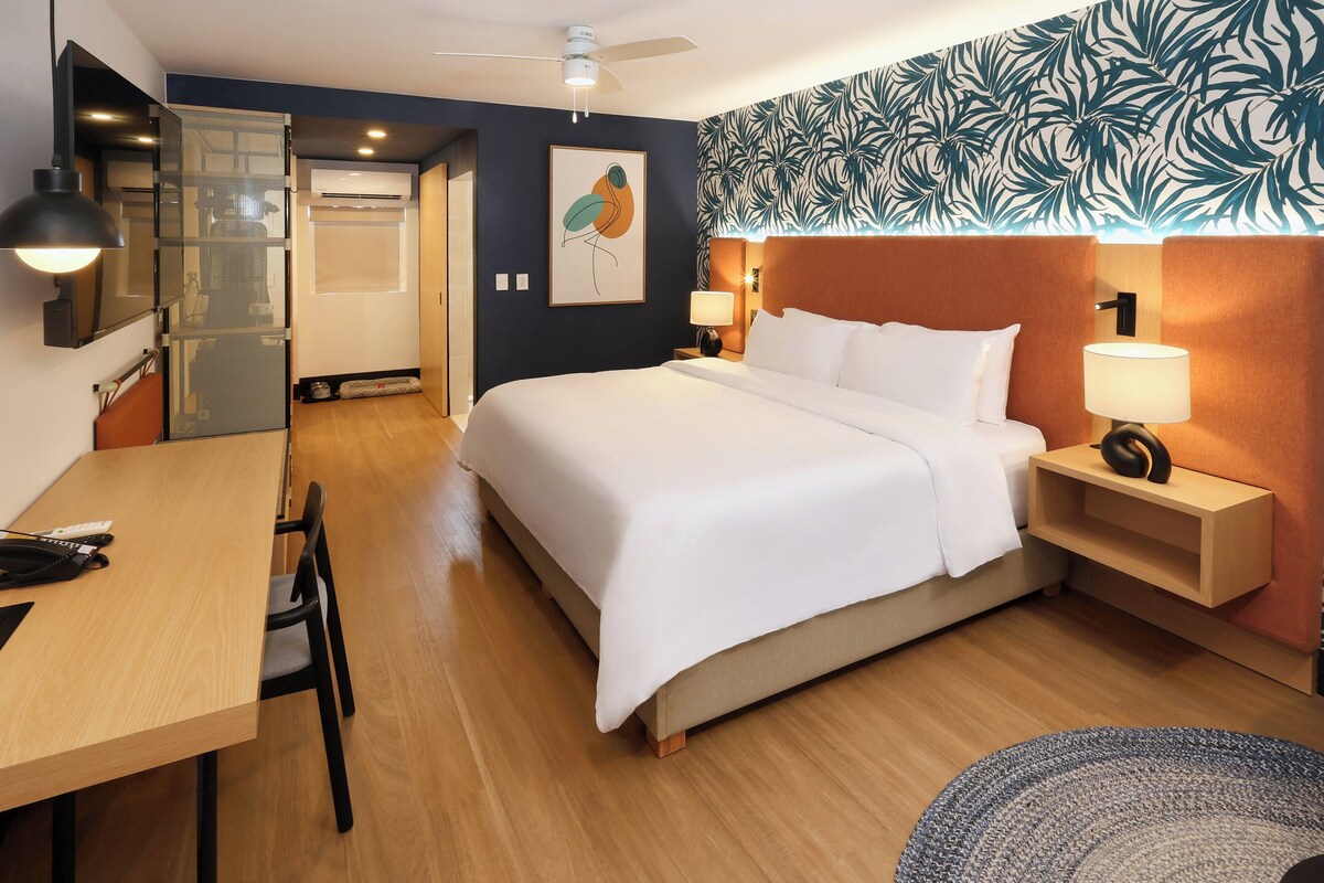 Habitación de hotel con cama, mesitas de noche, escritorio, silla, ventilador de techo y una pared con estampado de hojas.