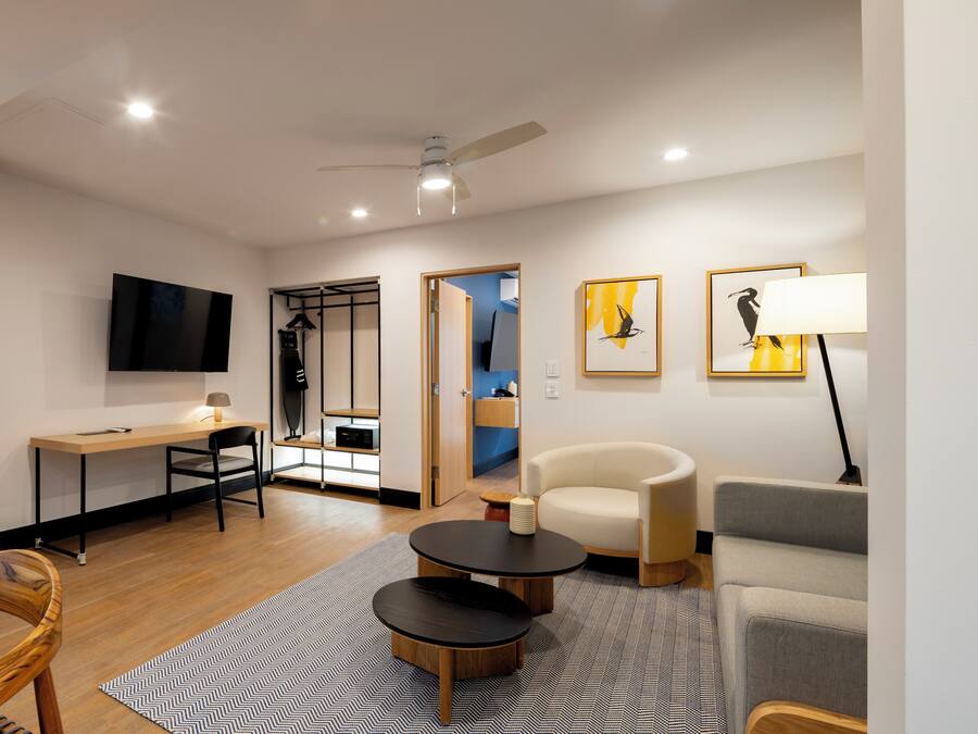 A modern living room with a flat-screen TV, a desk, a sofa, a chair, and a lamp.