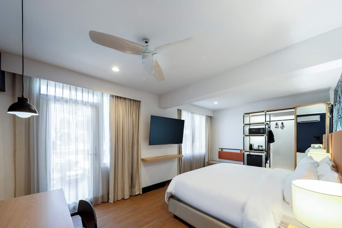 A hotel room with a bed, a desk, a chair, a TV mounted on the wall, a ceiling fan, and a minibar.