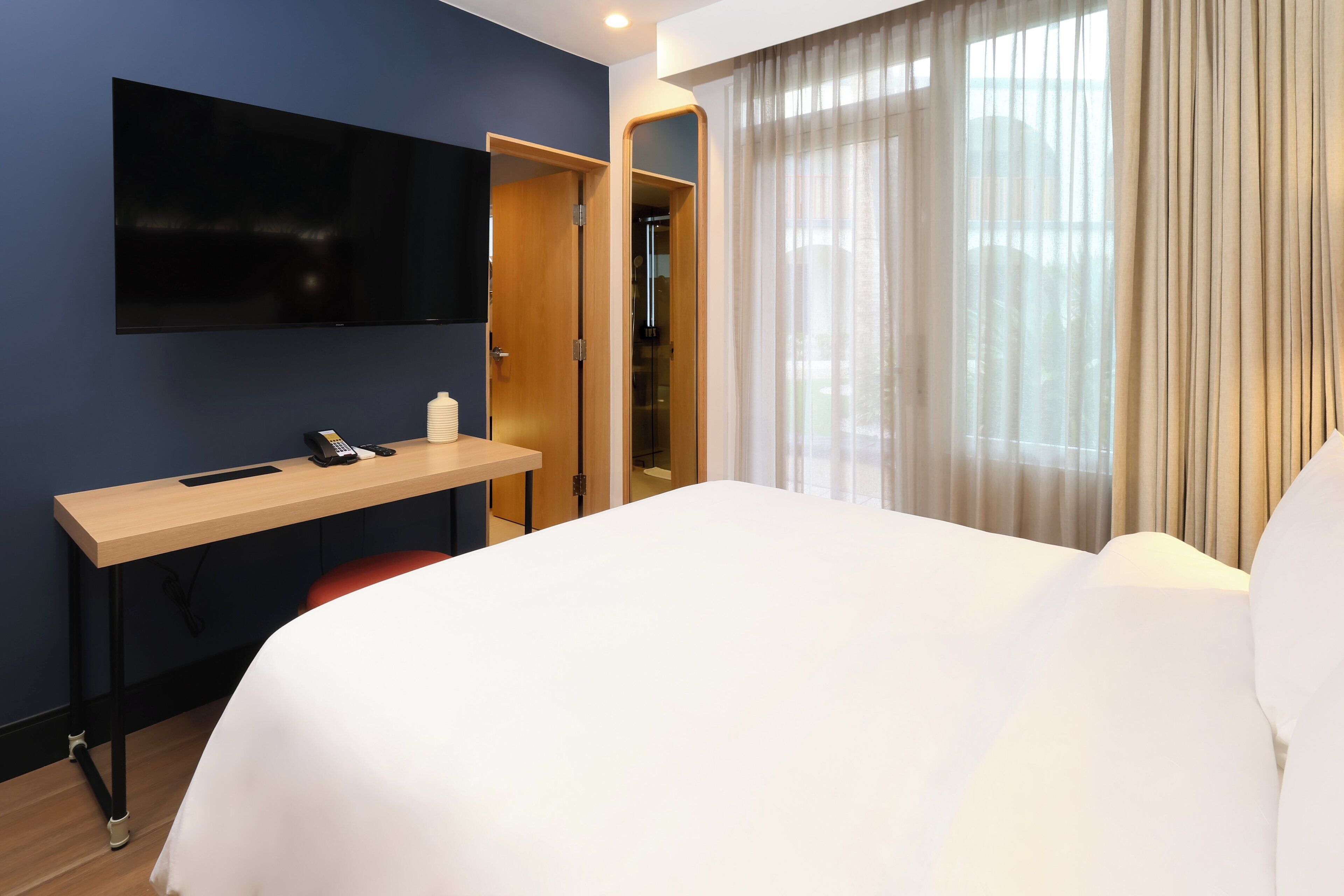 A hotel room with a bed, a desk with a phone, a TV mounted on the wall, and a window with curtains.