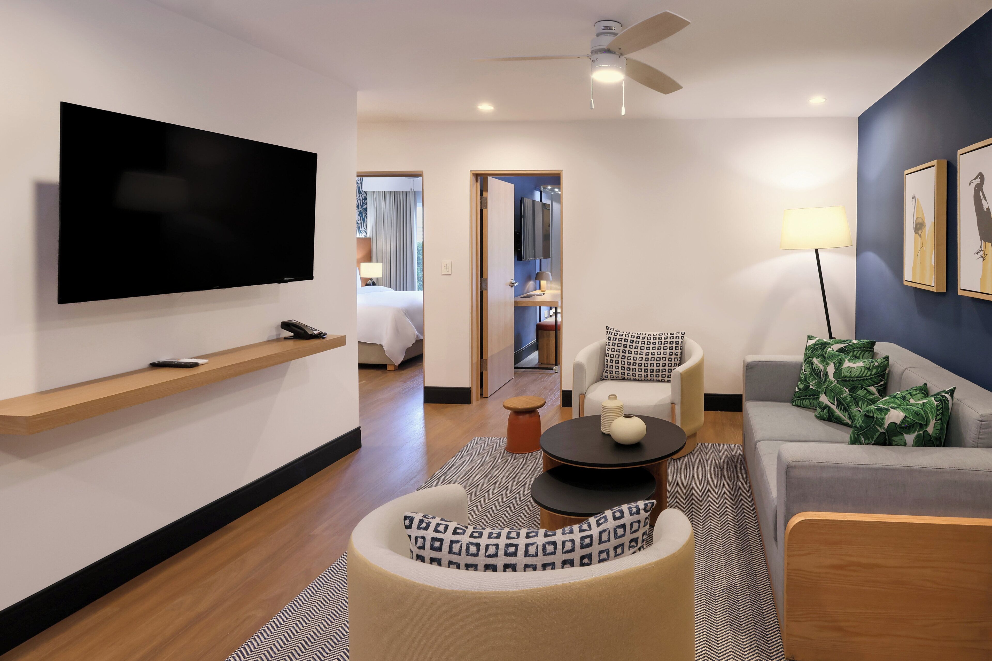 A modern hotel room with a flat-screen TV, a sofa, a chair, a small table, and a ceiling fan.