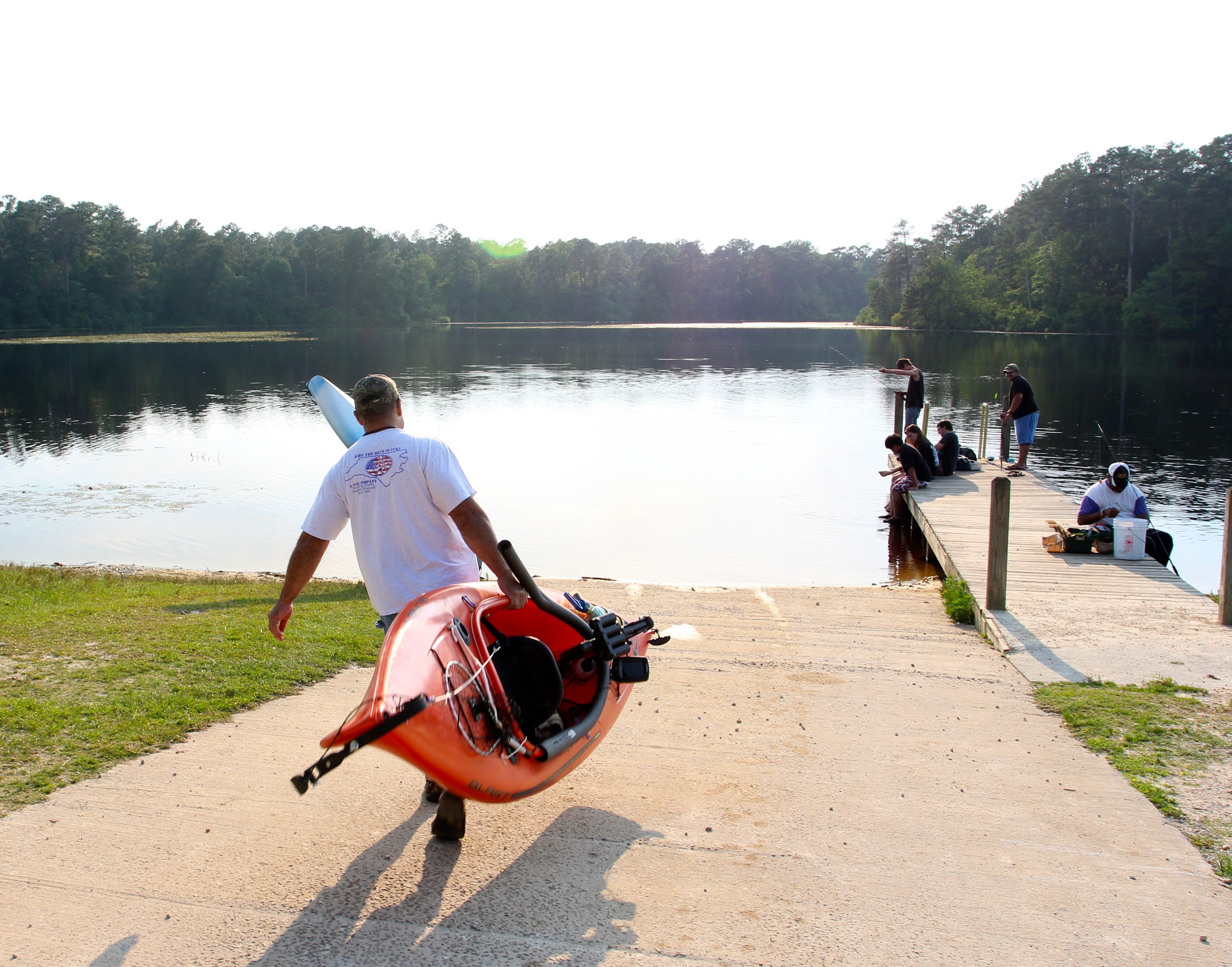 Um homem carregando um caiaque em um caminho de concreto ao lado de um lago calmo, com pessoas sentadas e em pé em um píer.