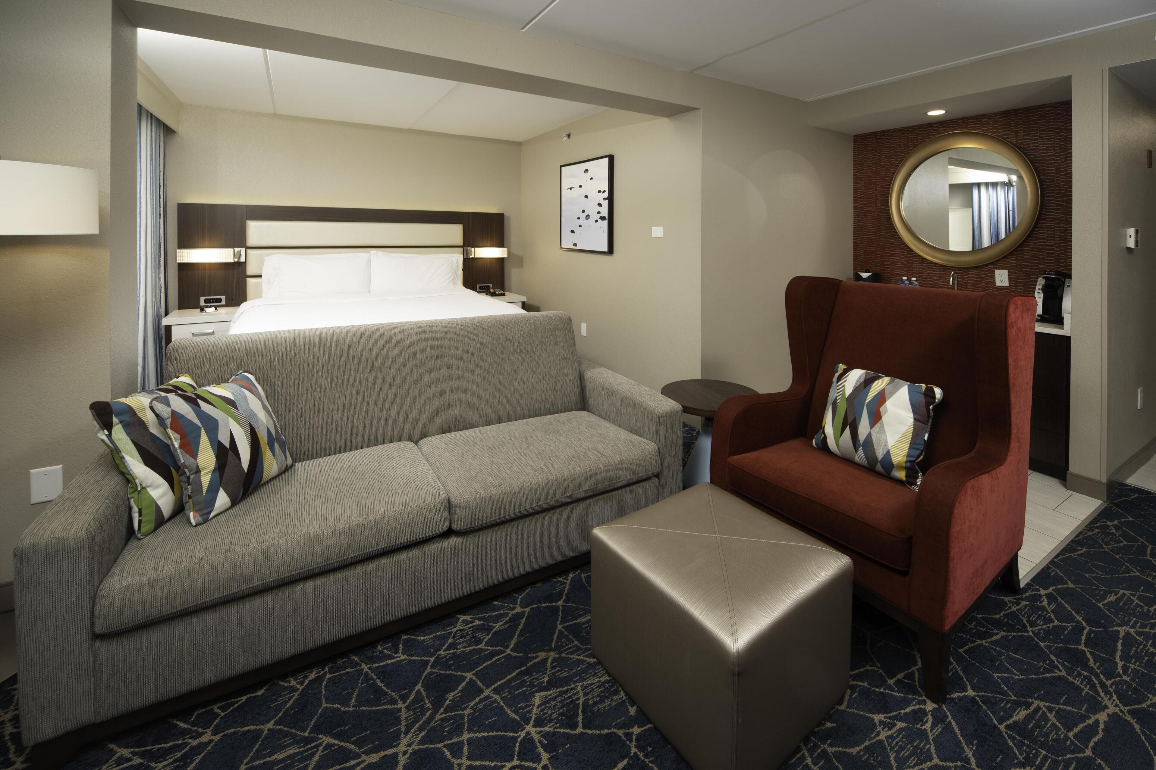 Une chambre d’hôtel avec un lit, un canapé gris, un fauteuil rouge et une petite table.