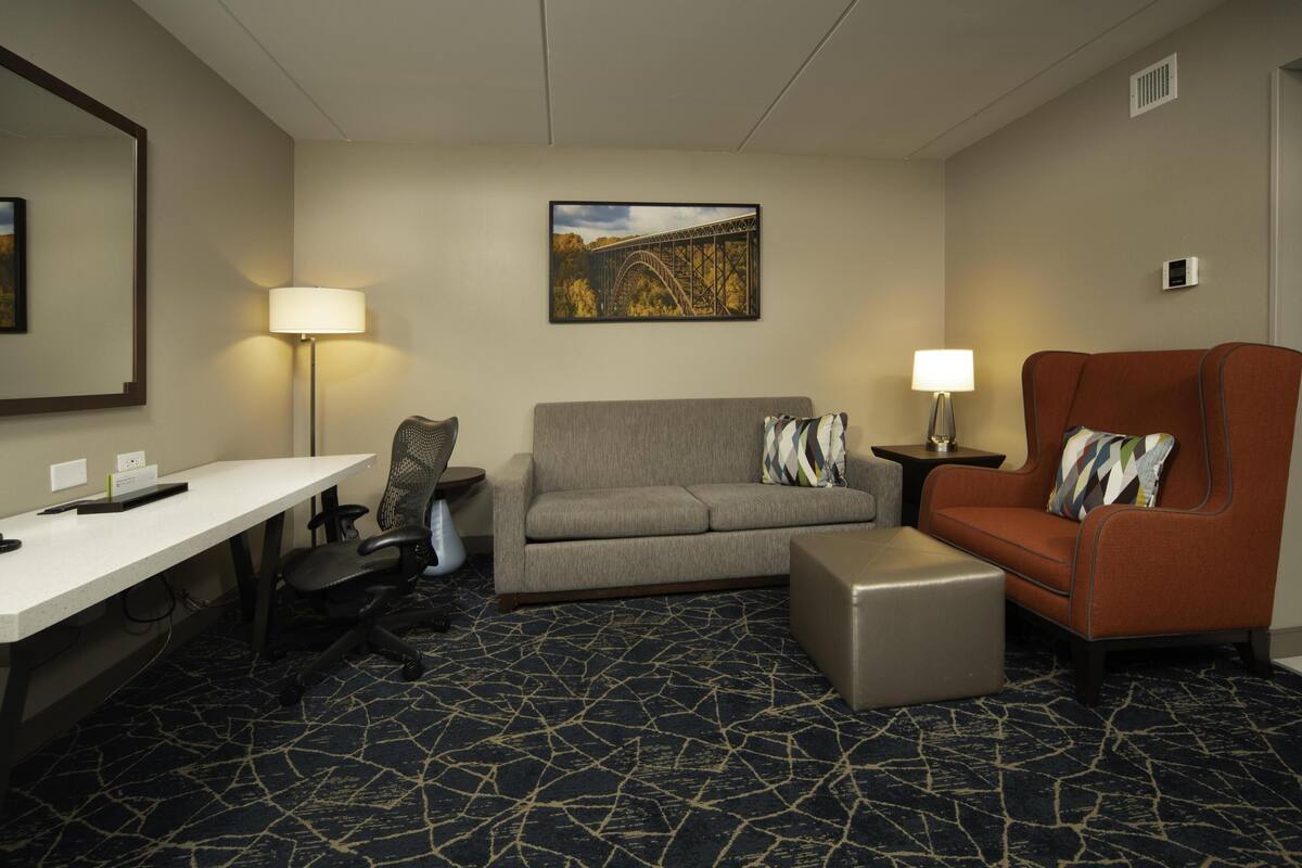 Una habitación de hotel con un escritorio, un sofá, un sillón, una mesita de centro, una lámpara y un cuadro en la pared.