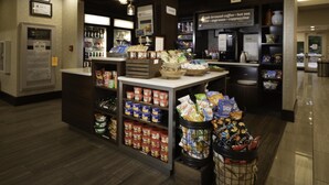 A coffee shop interior with a counter displaying various coffee beans and snacks. There are shelves stocked with packaged goods and a trash bin filled with snack bags.