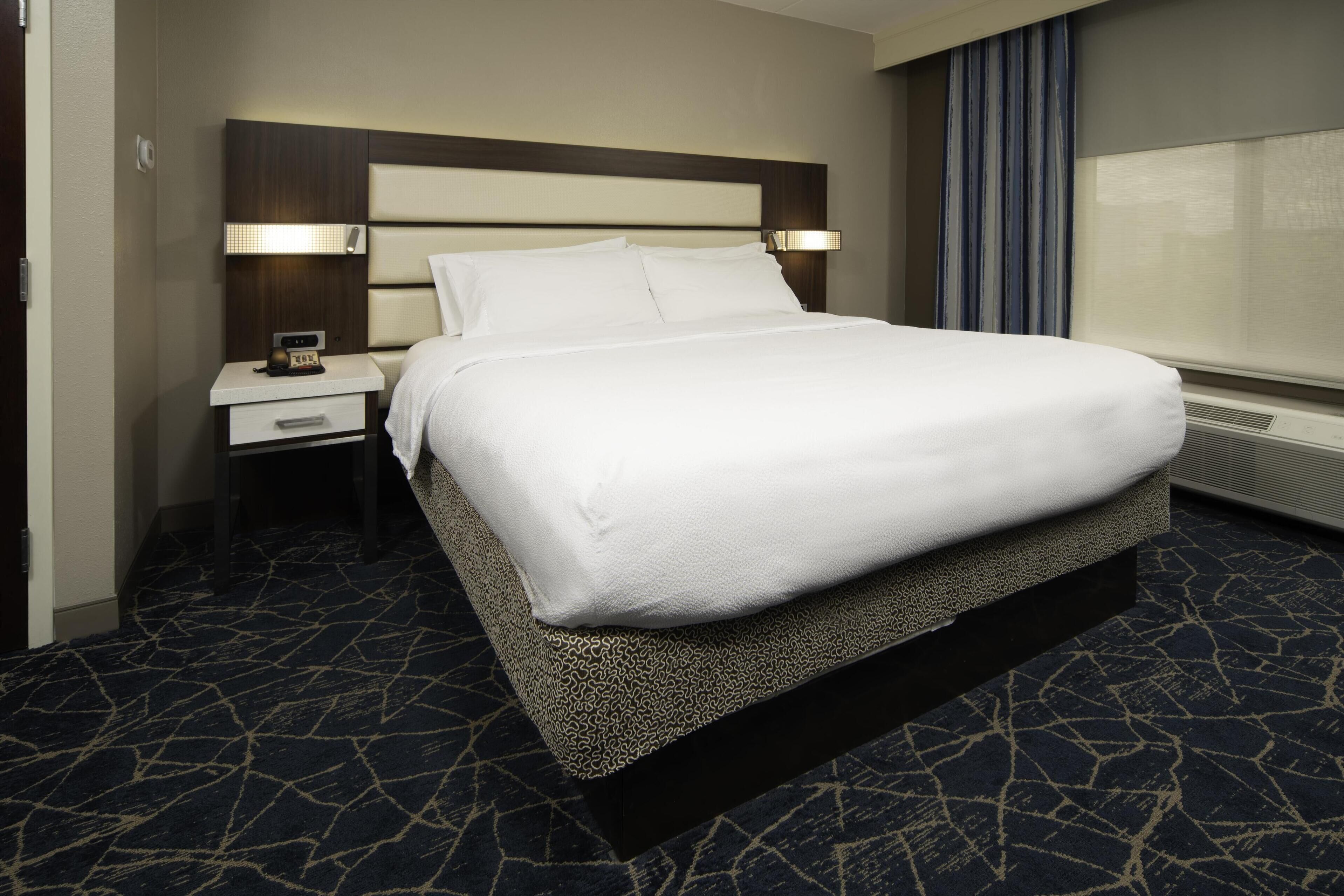 A hotel room with a large bed, a nightstand with a lamp, and a window with curtains.