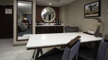 A modern hotel room with a dining area, a large mirror, a flat-screen TV, and a view of the lobby.