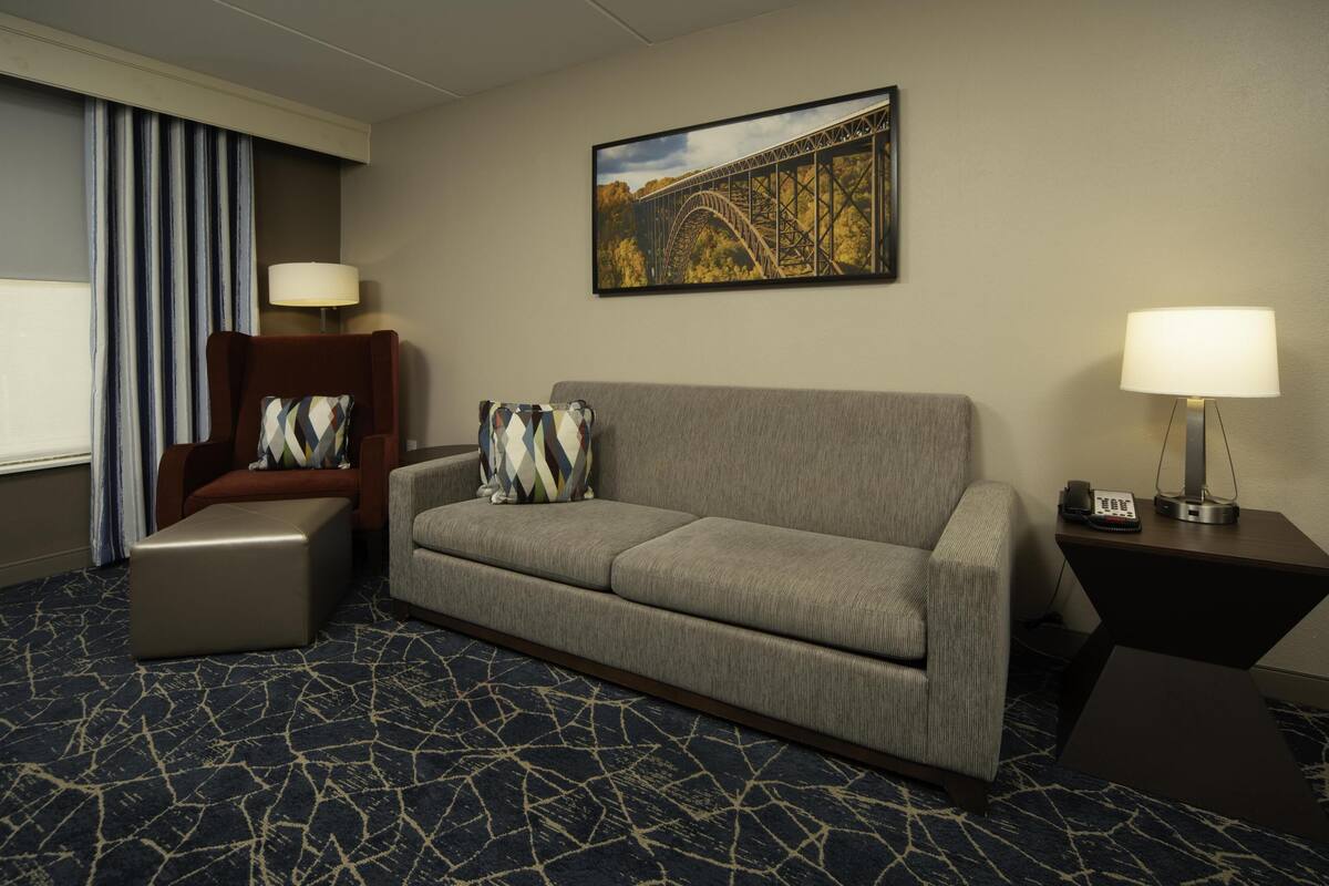Habitación de hotel con un sofá gris, un sillón marrón, un otoman estampado, un cuadro de un puente y una alfombra con diseño.