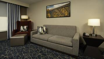 Habitación de hotel con un sofá gris, un sillón marrón, un otoman estampado, un cuadro de un puente y una alfombra con diseño.
