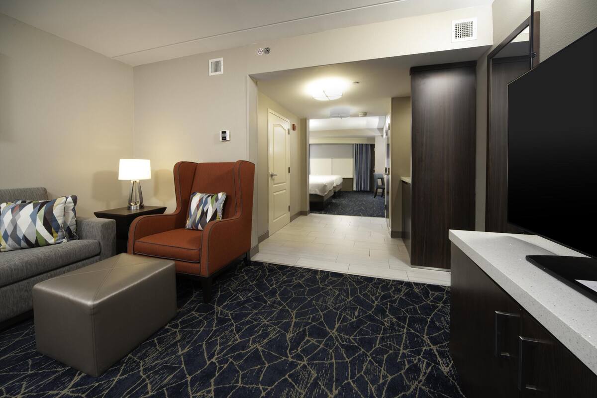 Habitación de hotel con sofá, sillón, otoman, televisor de pantalla plana, una pequeña mesa con lámpara y un área de kitchenette.