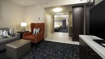 Une chambre d’hôtel équipée d’un canapé, d’un fauteuil, d’un pouf, d’un téléviseur à écran plat, d’une petite table avec une lampe et d’un coin cuisine.