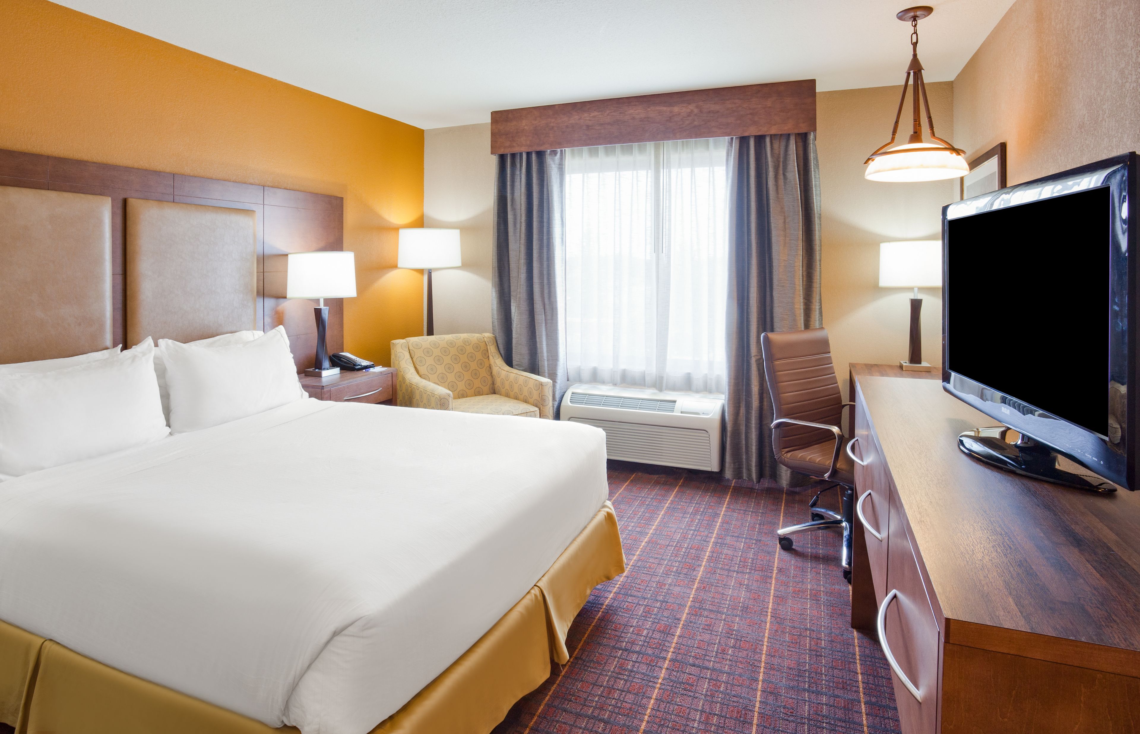 A hotel room with a large bed, a desk with a television, a chair, and a desk lamp.