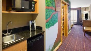 Habitación de hotel compacta con kitchenette que incluye microondas, pileta y lavavajillas. Un mural con un árbol y césped decora la pared.
