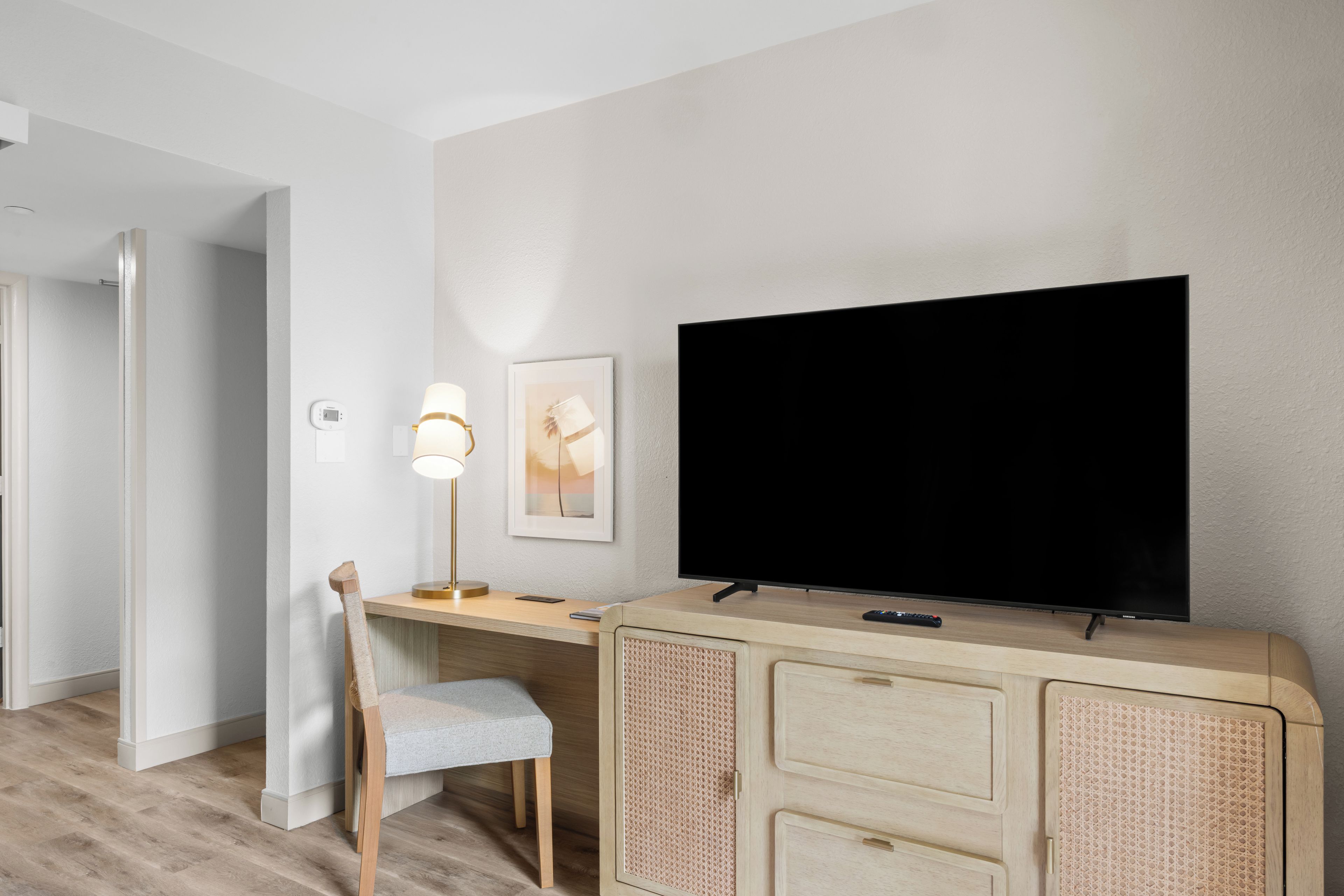 Een moderne kamer met een flatscreen-televisie aan de muur, een bureau met een lamp, een stoel en een rotan opbergmeubel.