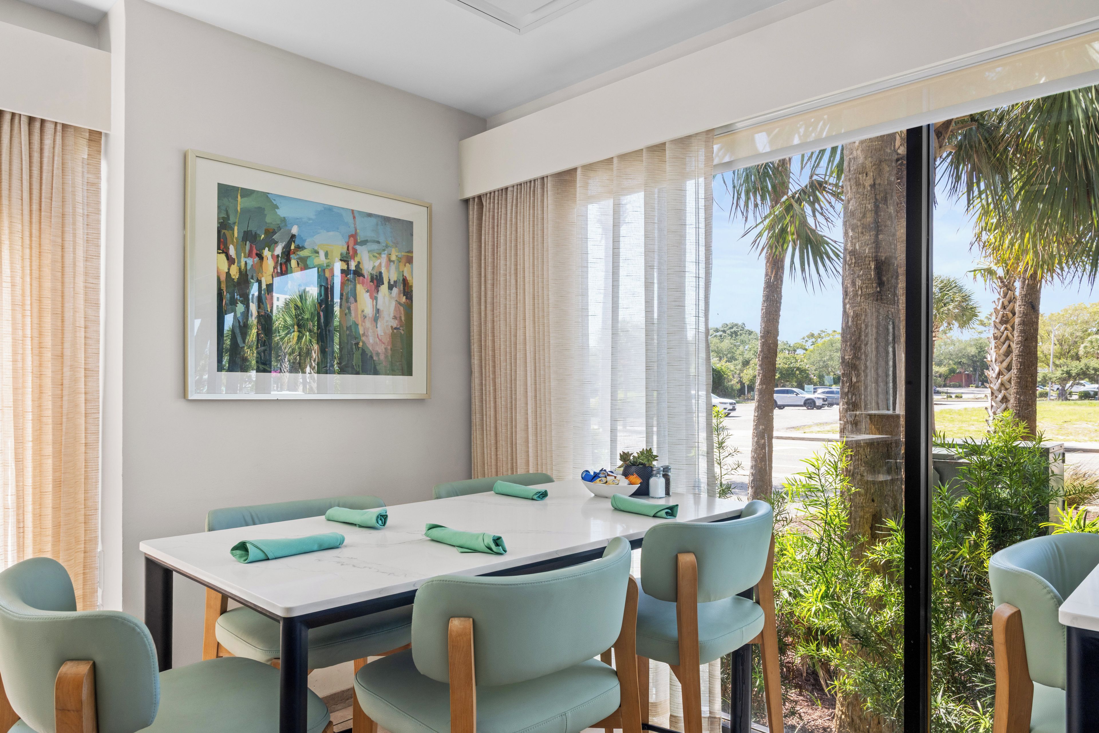 A dining area with a table, chairs, a painting, and a view of palm trees.