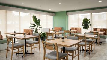 A modern indoor dining area with wooden tables and chairs, potted plants, and large windows.