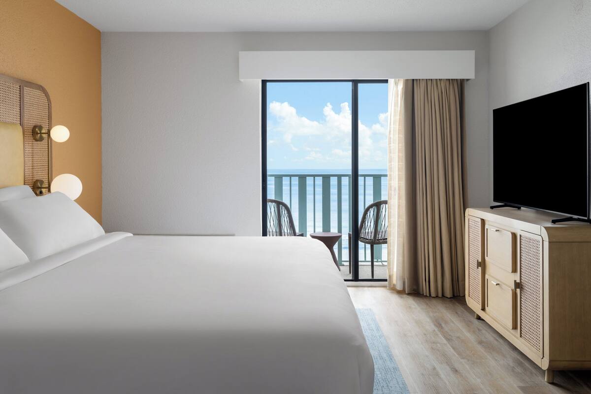 A hotel room with a large bed, a television, a small table, and a balcony with a view of the sea.