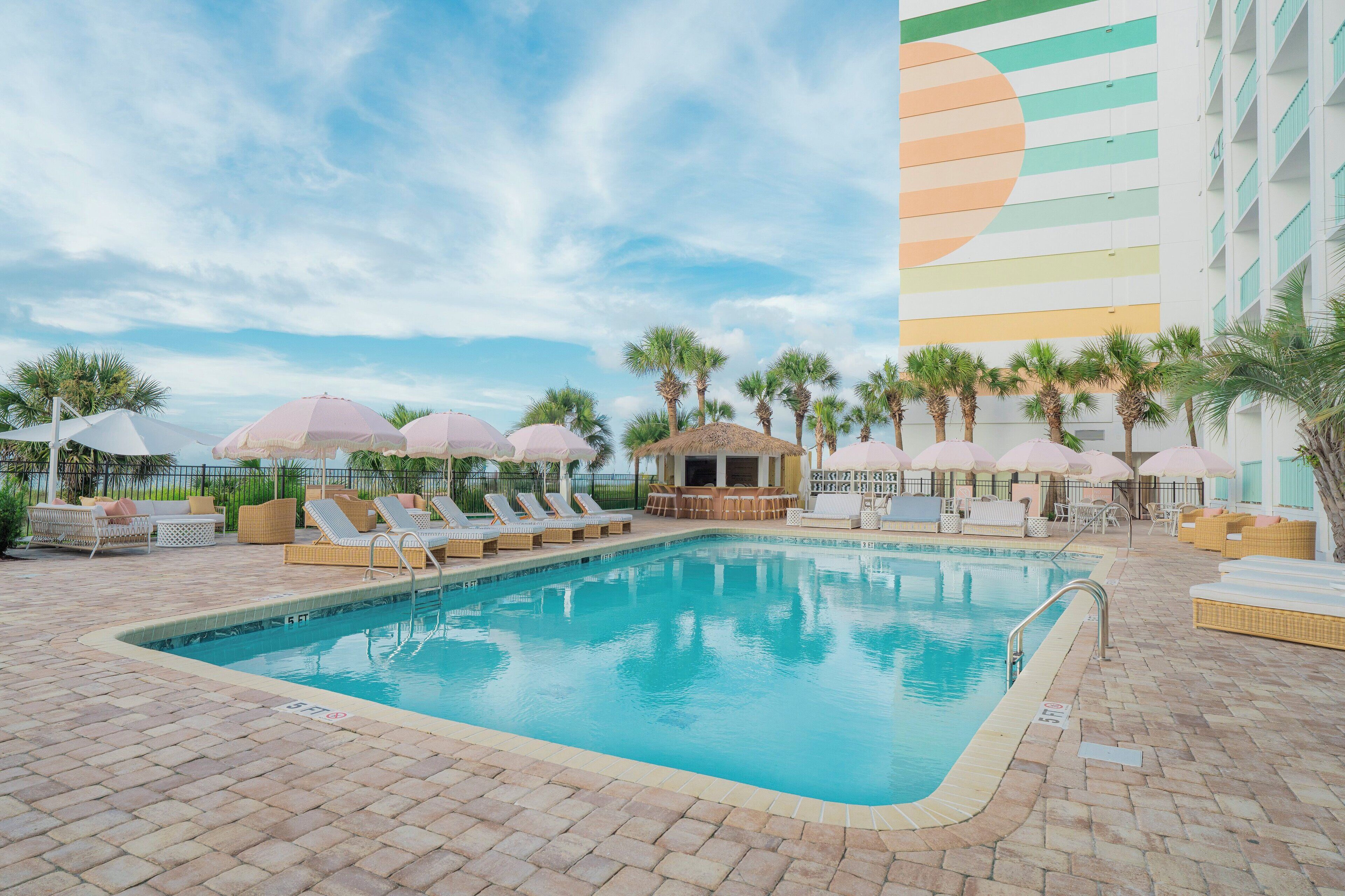 A swimming pool with lounge chairs and umbrellas, surrounded by palm trees and a large building with a colorful mural.
