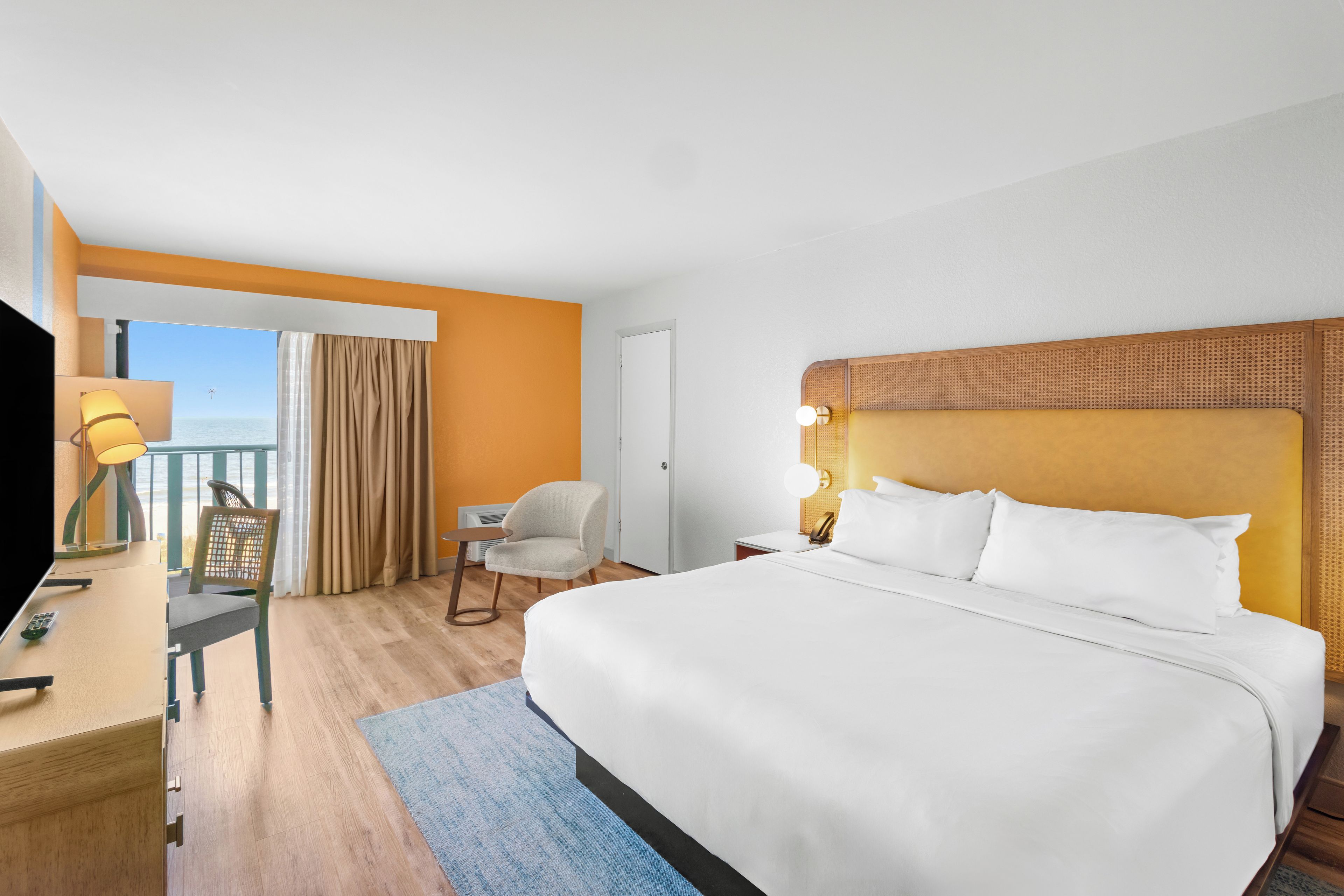 Quarto de hotel com uma cama grande, uma escrivaninha com abajur, uma cadeira e vista para o oceano.
