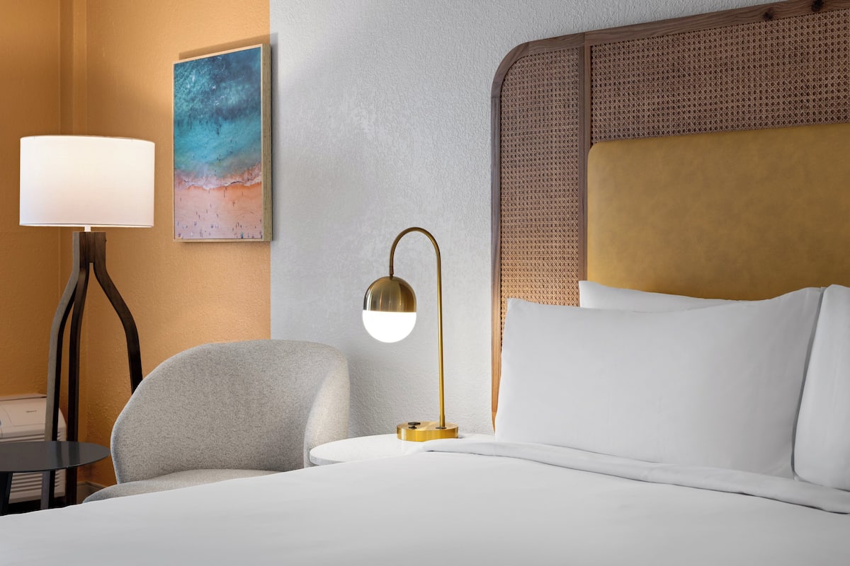 A hotel room with a bed, a lamp, a chair, and a painting on the wall.